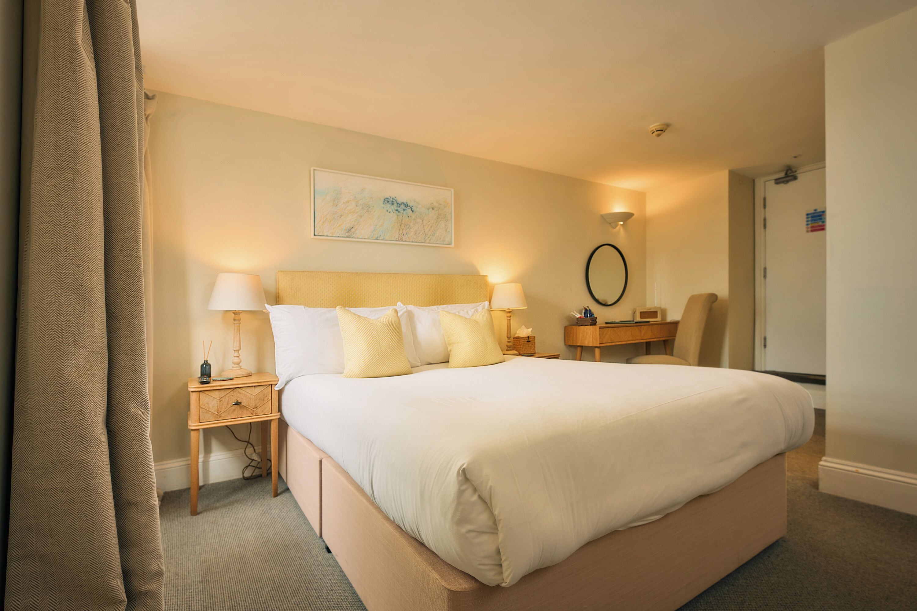 A hotel room with a bed, bedside lamps, a desk, a chair, a mirror, and a door.