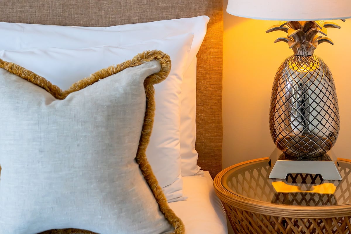 A neatly made bed with a white pillow and a textured pillow with a golden trim. A lamp with a pineapple-shaped base is on a wicker side table beside the bed.