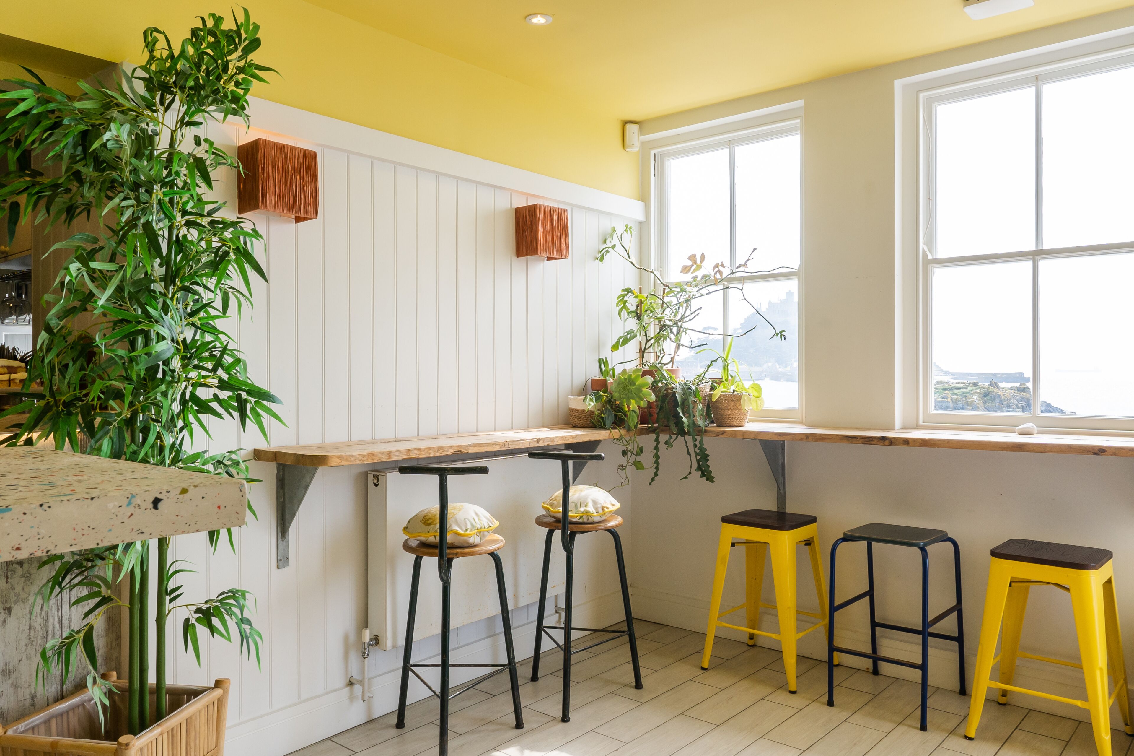 Cozinha com painéis de madeira branca, teto amarelo e janela com vista. Há três banquetas e uma planta no balcão.