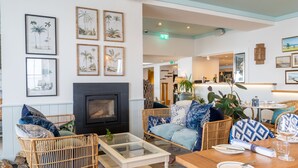 A cozy lounge area with wicker furniture, a fireplace, and tropical-themed wall art. - The Godolphin (Marazion)