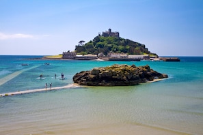 A coastal scene with a prominent castle on a hill, clear turquoise waters, and a rocky outcrop in the foreground. - The Godolphin (Marazion)