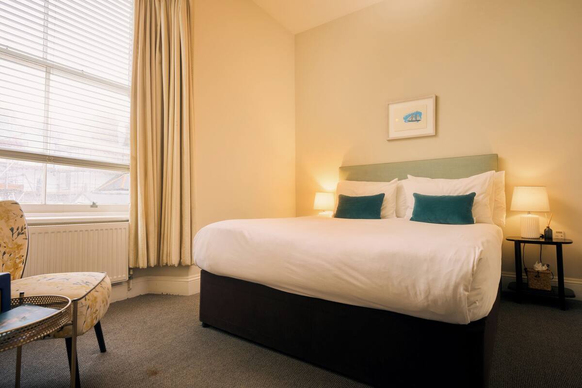 A hotel room with a bed, two bedside lamps, a chair, a window with curtains, and a picture on the wall.
