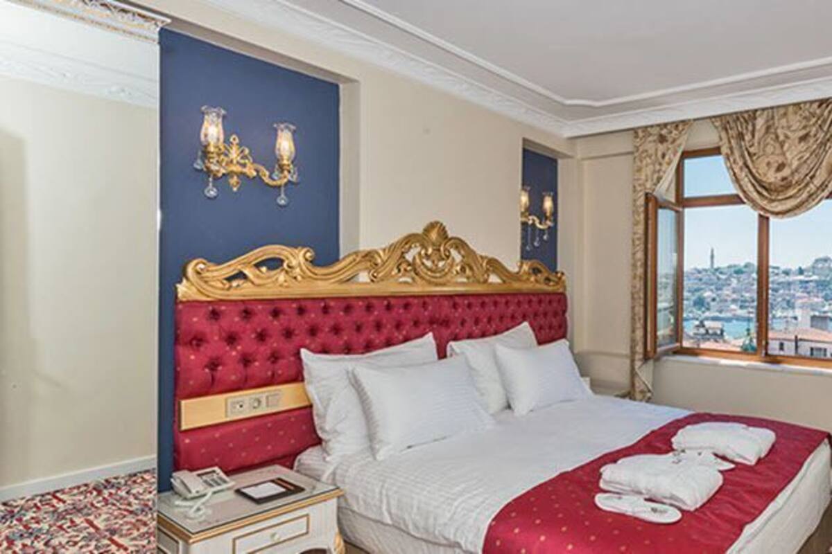 A hotel room with a large bed, a nightstand, a TV, and a view of a cityscape through the window.