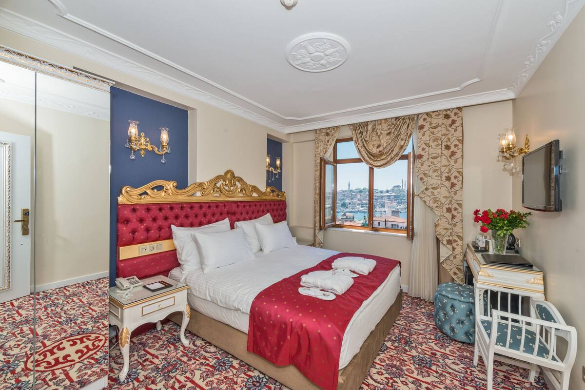 A hotel room with a large bed, a nightstand, a small table, a crib, a TV, and a window with a city view.