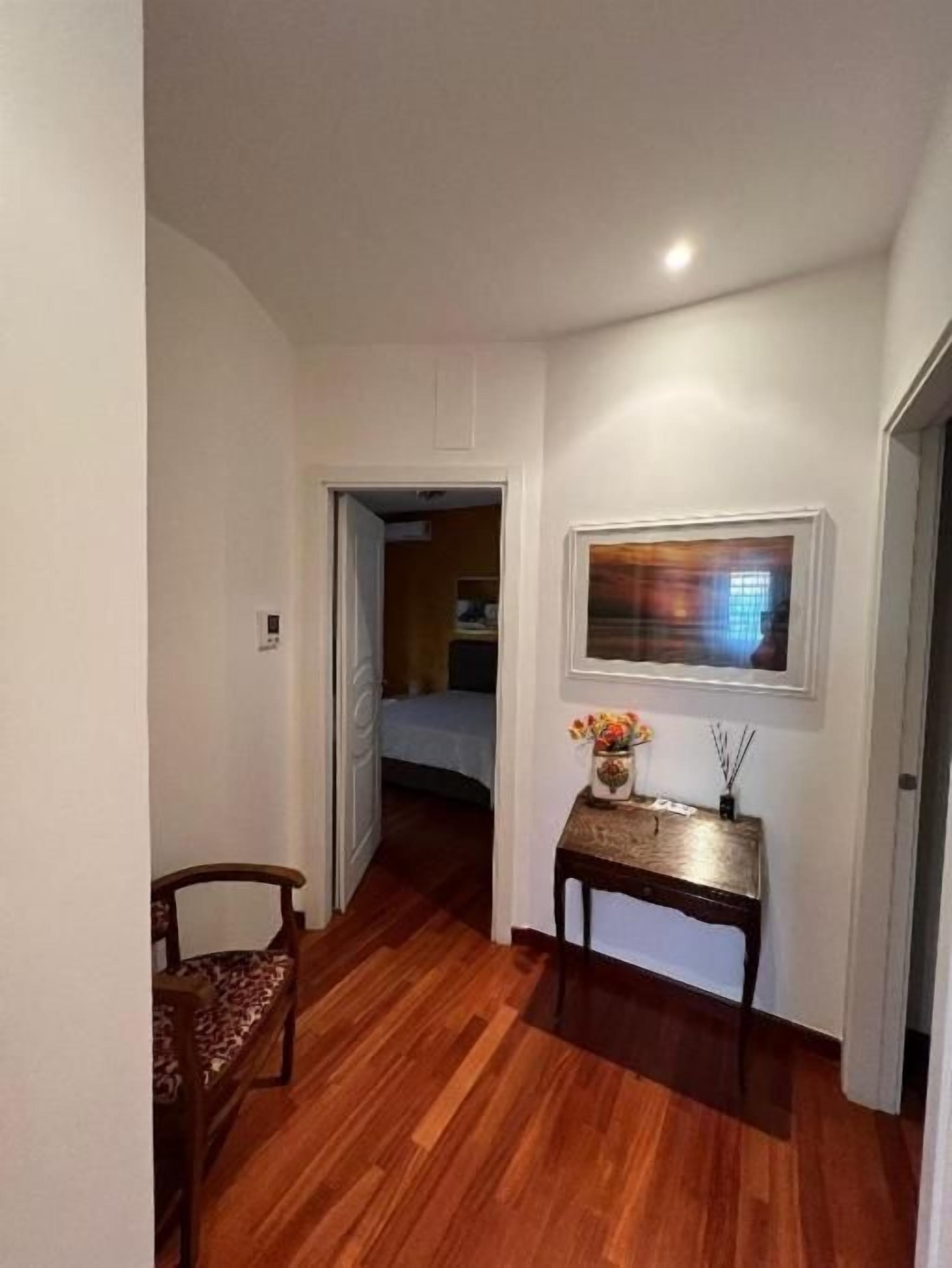 A hallway with wooden flooring, a door leading to a bedroom, a small table with a vase, and a framed picture on the wall.