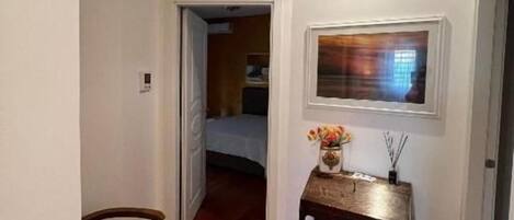 A hallway with wooden flooring, a door leading to a bedroom, a small table with a vase, and a framed picture on the wall.