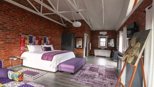 Un dormitorio con pared de ladrillo, cama con ropa de cama blanca y morada, suelo de madera y un cuadro en la pared.