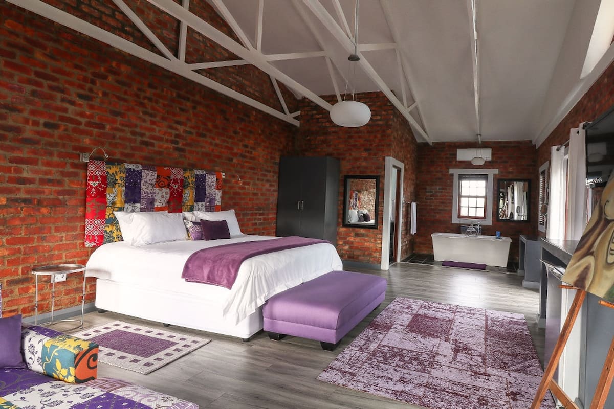 Une chambre avec un mur en briques, un lit recouvert d’une literie blanche et violette, un plancher en bois et un tableau accroché au mur.