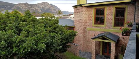 A brick house with a green roof and yellow trim, surrounded by lush greenery and mountains in the background.