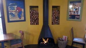 Una habitación acogedora con chimenea, sillas de madera y una mesa. Hay dos estantes en la pared con leña y un cuadro enmarcado.