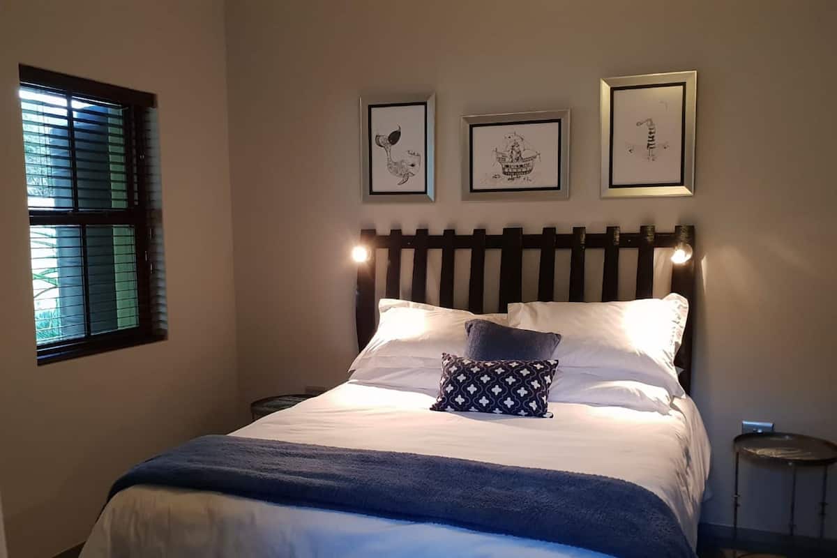 Un lit bien fait, avec une literie blanche et bleue, un coussin à motifs et trois œuvres d’art encadrées accrochées au mur.