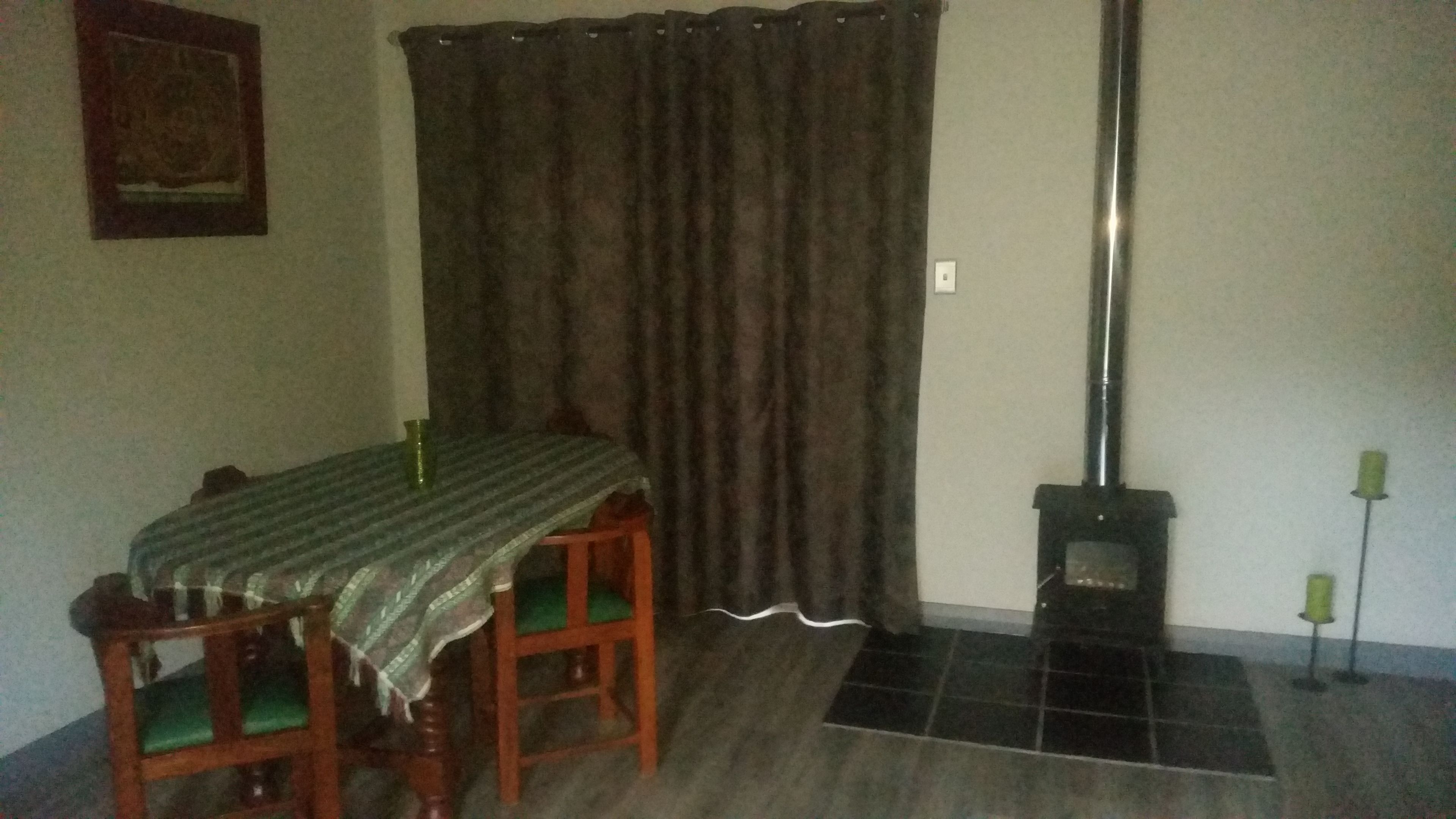 A dining area with a table covered by a tablecloth, two chairs, a curtain, a fireplace, and a framed picture on the wall.
