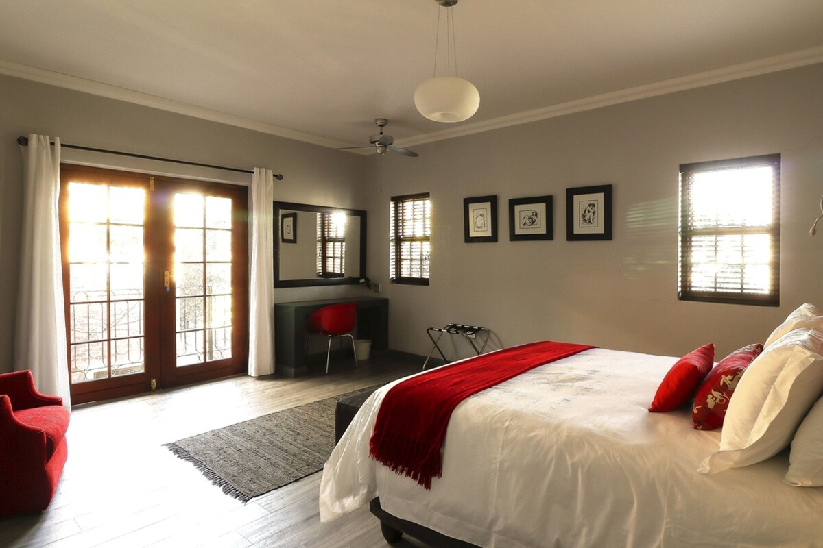 A bedroom with a large bed, a desk, and a chair. There are windows with blinds, a ceiling fan, and framed pictures on the wall.