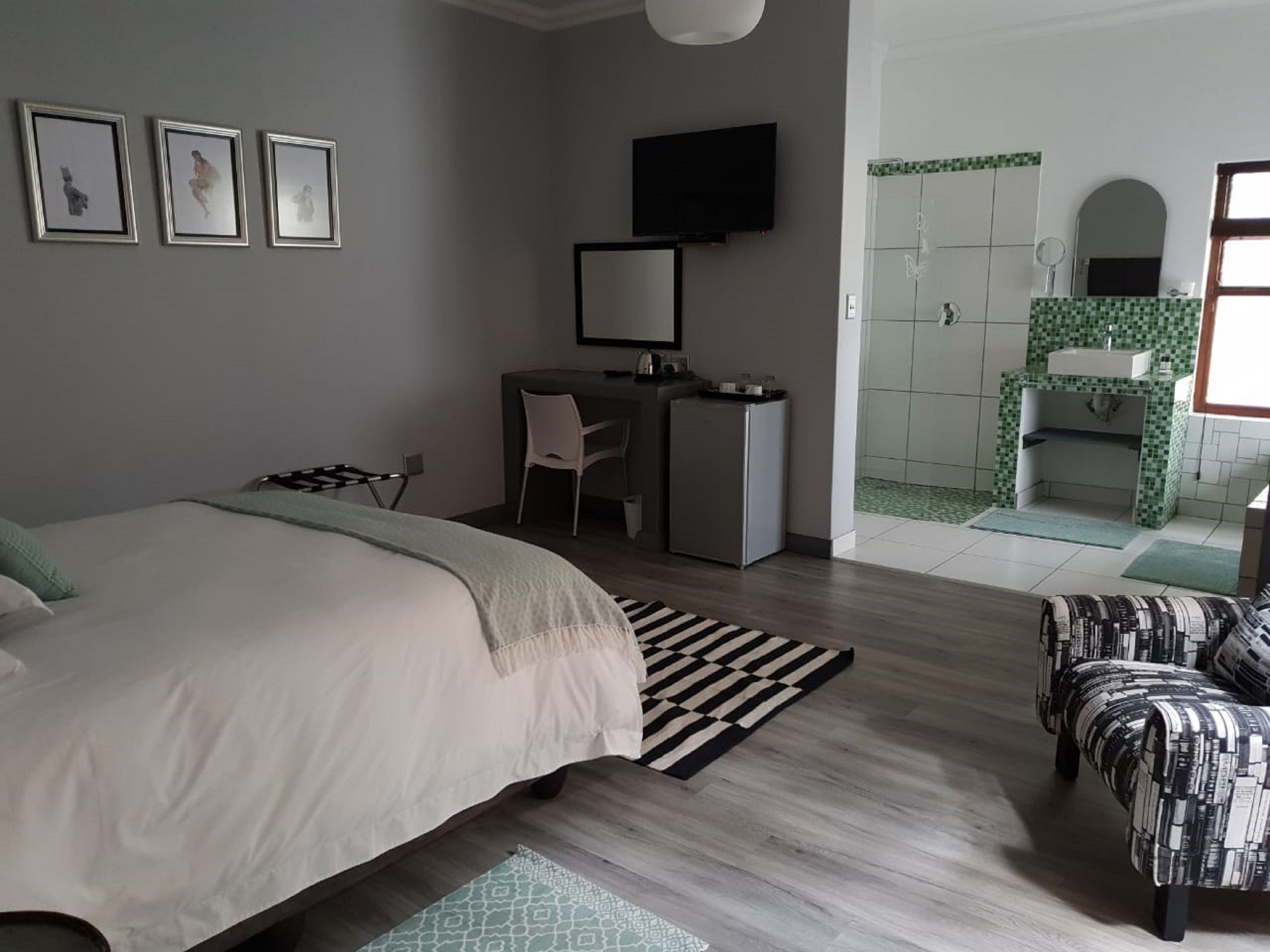 Quarto com cama, escrivaninha, cadeira, televisão e banheiro com chuveiro e pia.