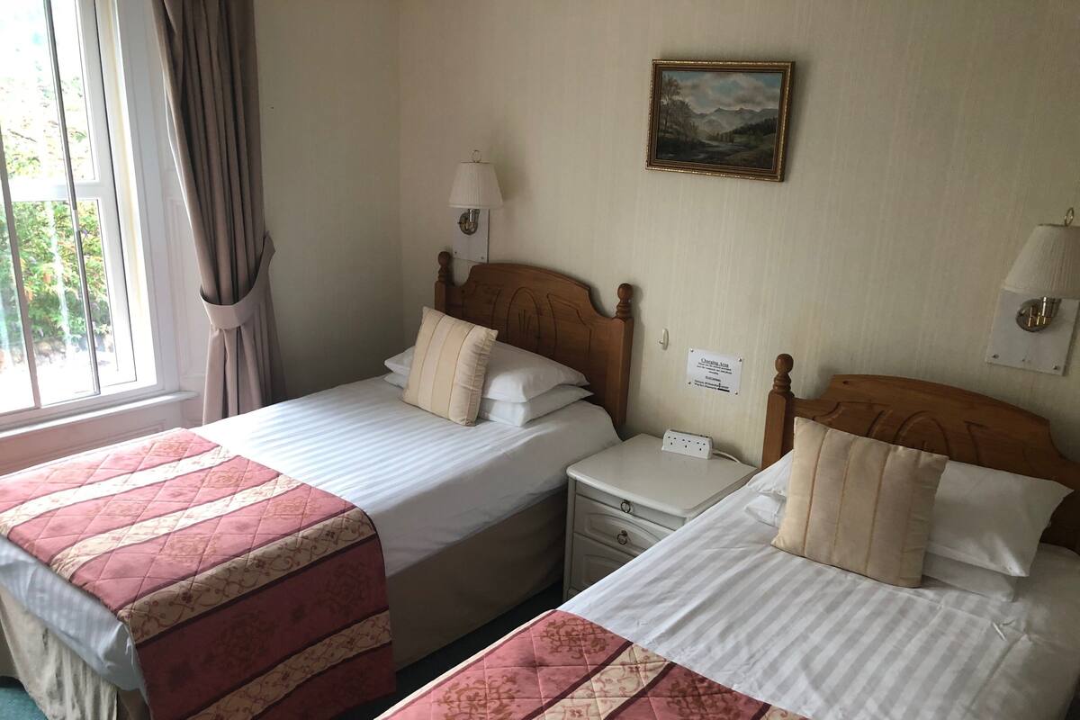 A hotel room with two beds, each with a red and beige quilted bedspread, a wooden headboard, a nightstand with a lamp, and a wall-mounted painting.