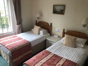 A hotel room with two beds, each with a red and beige quilted bedspread, a wooden headboard, a nightstand with a lamp, and a wall-mounted painting. - The Well House (Pitlochry)