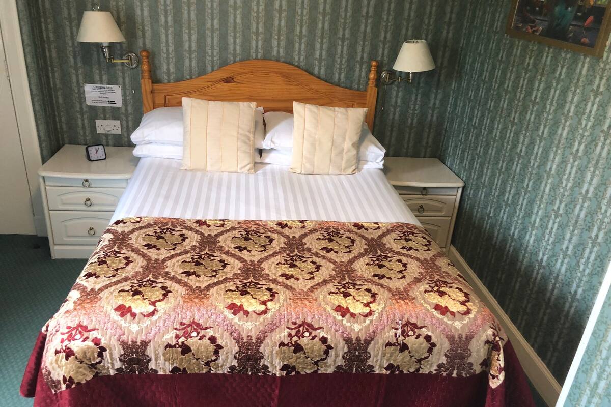 A neatly made bed with a patterned bedspread and two pillows, flanked by two bedside tables with lamps, in a room with striped wallpaper.