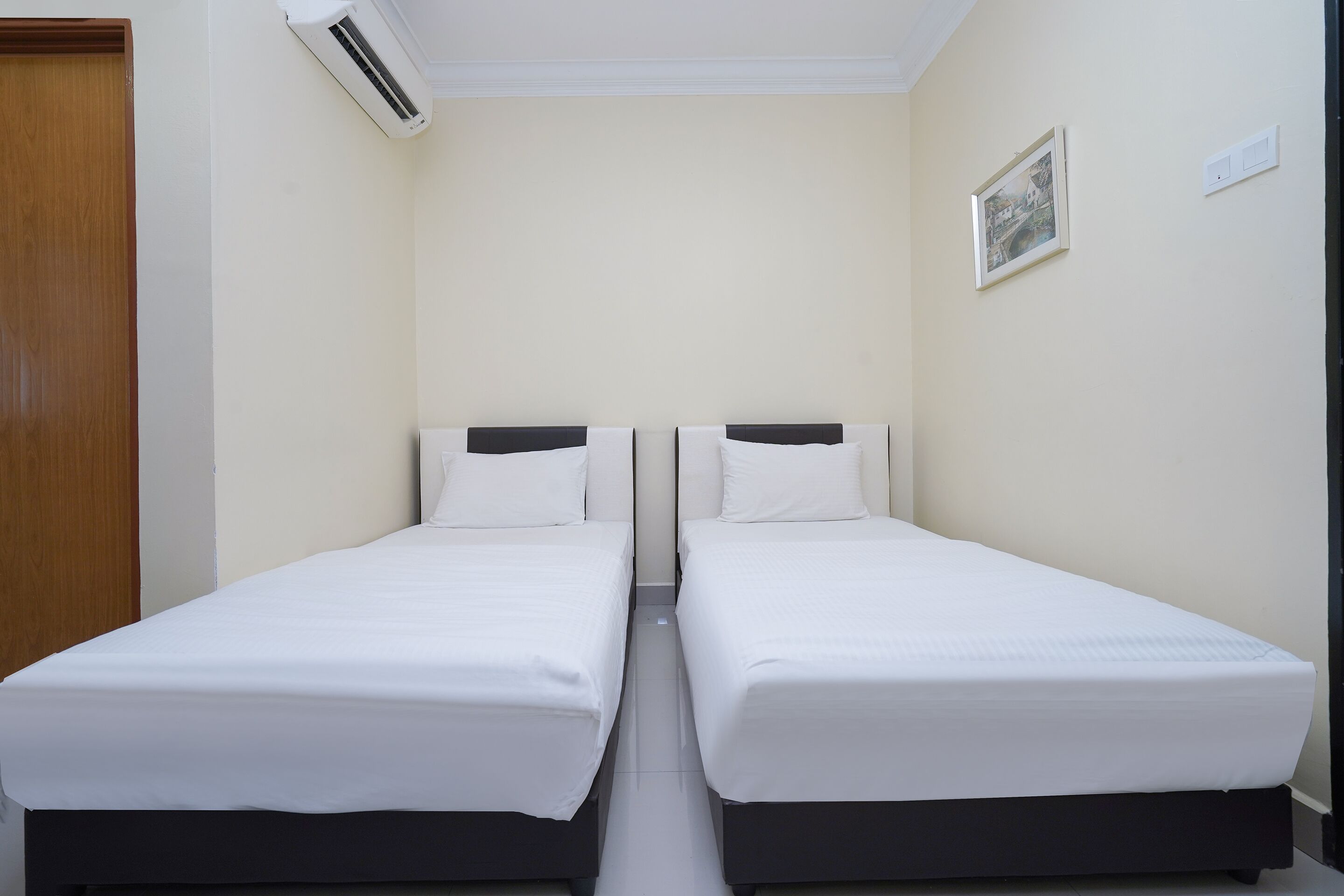 A room with two single beds, a wooden door, an air conditioning unit, a light switch, and a framed picture on the wall.