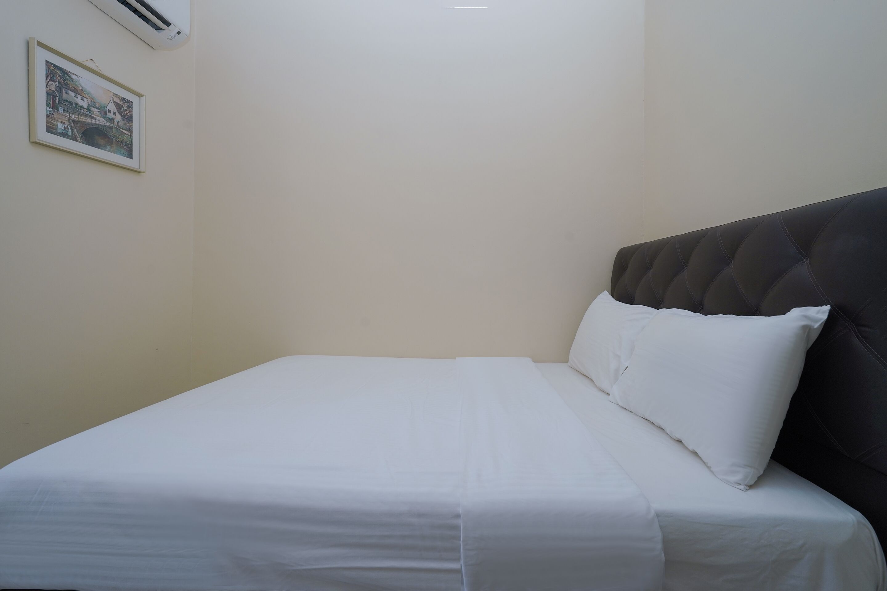 Un lit bien fait, avec du linge de lit blanc, dans une chambre dont la tête de lit est de couleur sombre et où un tableau encadré est accroché au mur.