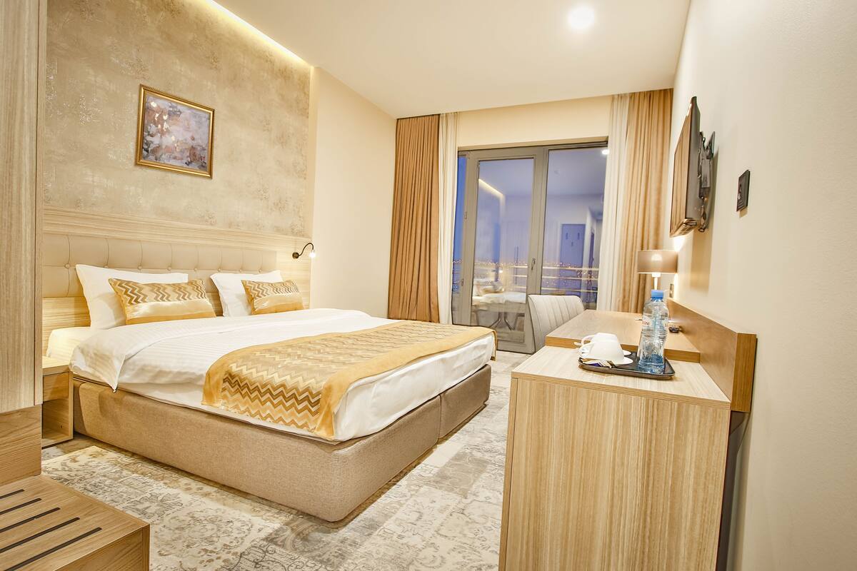 Habitación de hotel con una cama grande, un escritorio con lámpara, una botella de agua y vista a la ciudad por la ventana.