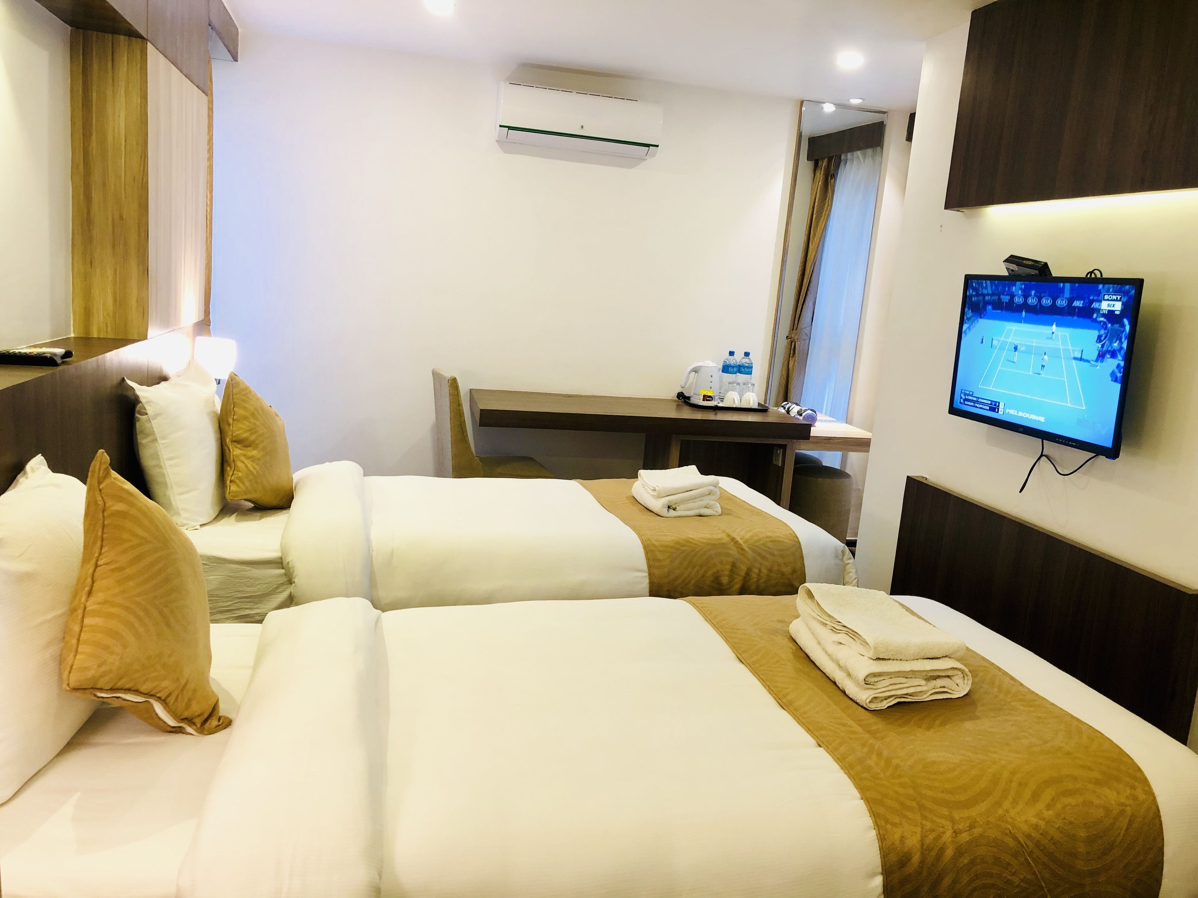A hotel room with two beds, a desk, a TV, and an air conditioning unit.