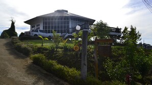 Un edificio moderno con un amplio frente de vidrio, rodeado de abundante vegetación y un sendero de tierra que conduce a la entrada.