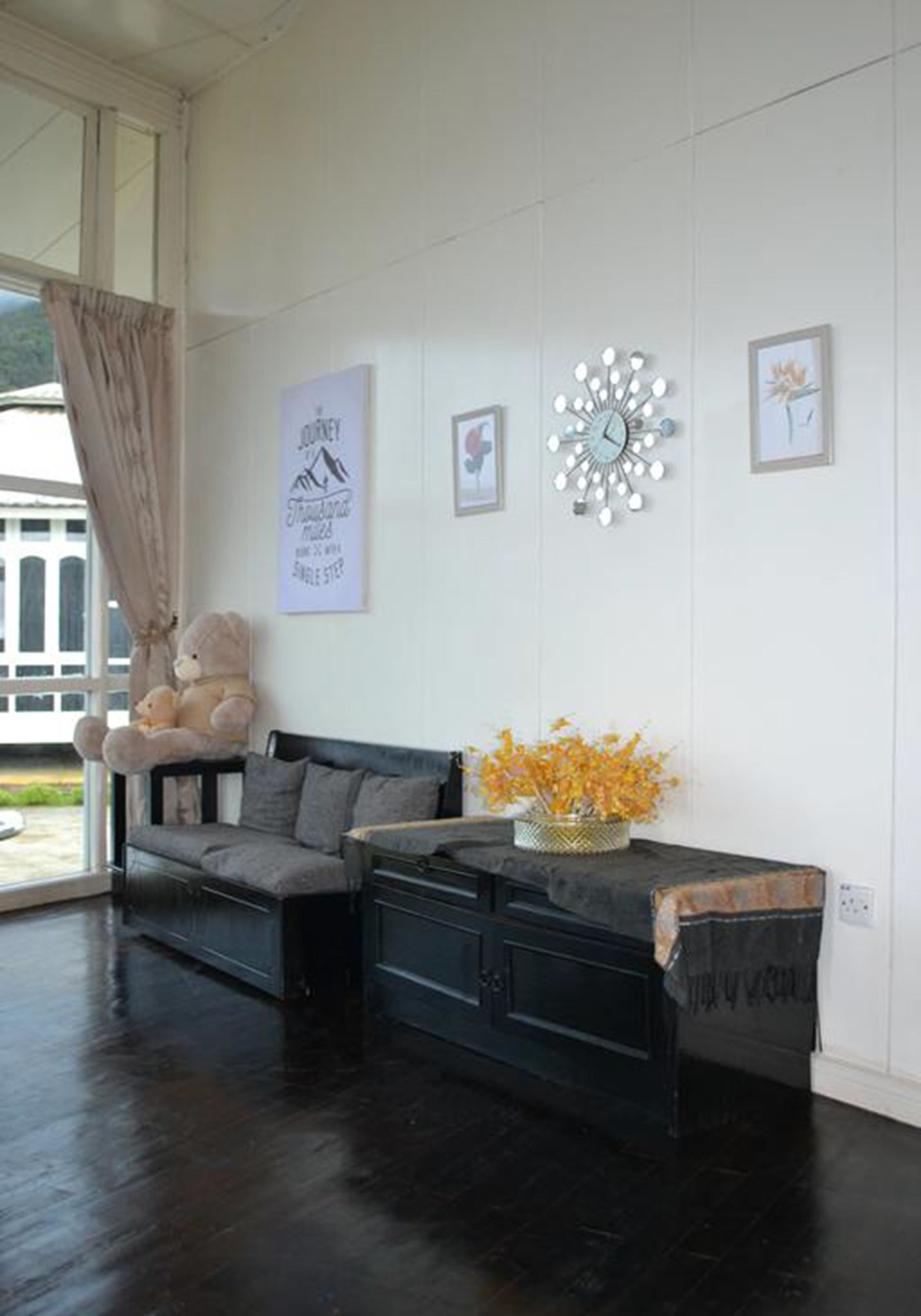 Une pièce avec un plancher en bois foncé, un canapé gris, une petite table sur laquelle est posé un vase de fleurs jaunes, et un ours en peluche sur le canapé.