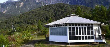 A small, single-story building with a tiled roof and large windows, surrounded by lush greenery and mountains in the background.