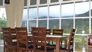 Una mesa de comedor de madera con seis sillas, amplias ventanas que ofrecen vistas a una montaña boscosa y una lámpara colgante.
