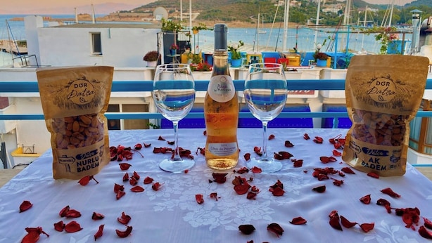 Une table avec deux sachets d’amandes, une bouteille de vin et deux verres, le tout posé sur une nappe parsemée de pétales de rose, avec vue sur une marina où sont amarrés des bateaux et sur des collines.