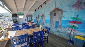 Un restaurant avec des chaises bleues et une table en bois, une fresque représentant des tours et la mer, et un panneau indiquant les directions.