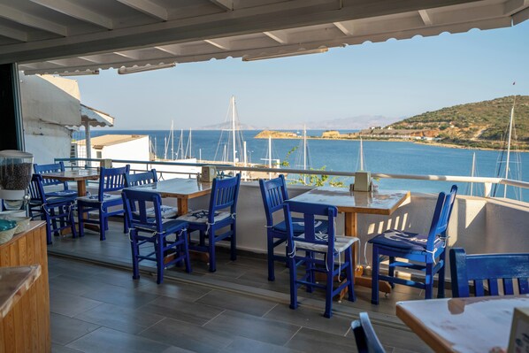 A covered outdoor seating area with blue chairs and tables, overlooking a marina with sailboats and a hilly coastline. - Sakin Otel (Datça)