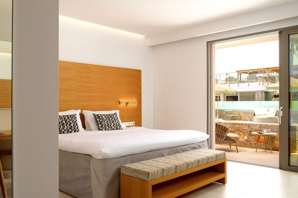 A modern hotel room with a wooden headboard, a large bed with white bedding, a bench, a desk with a chair, and a view of an outdoor area with tables and chairs.