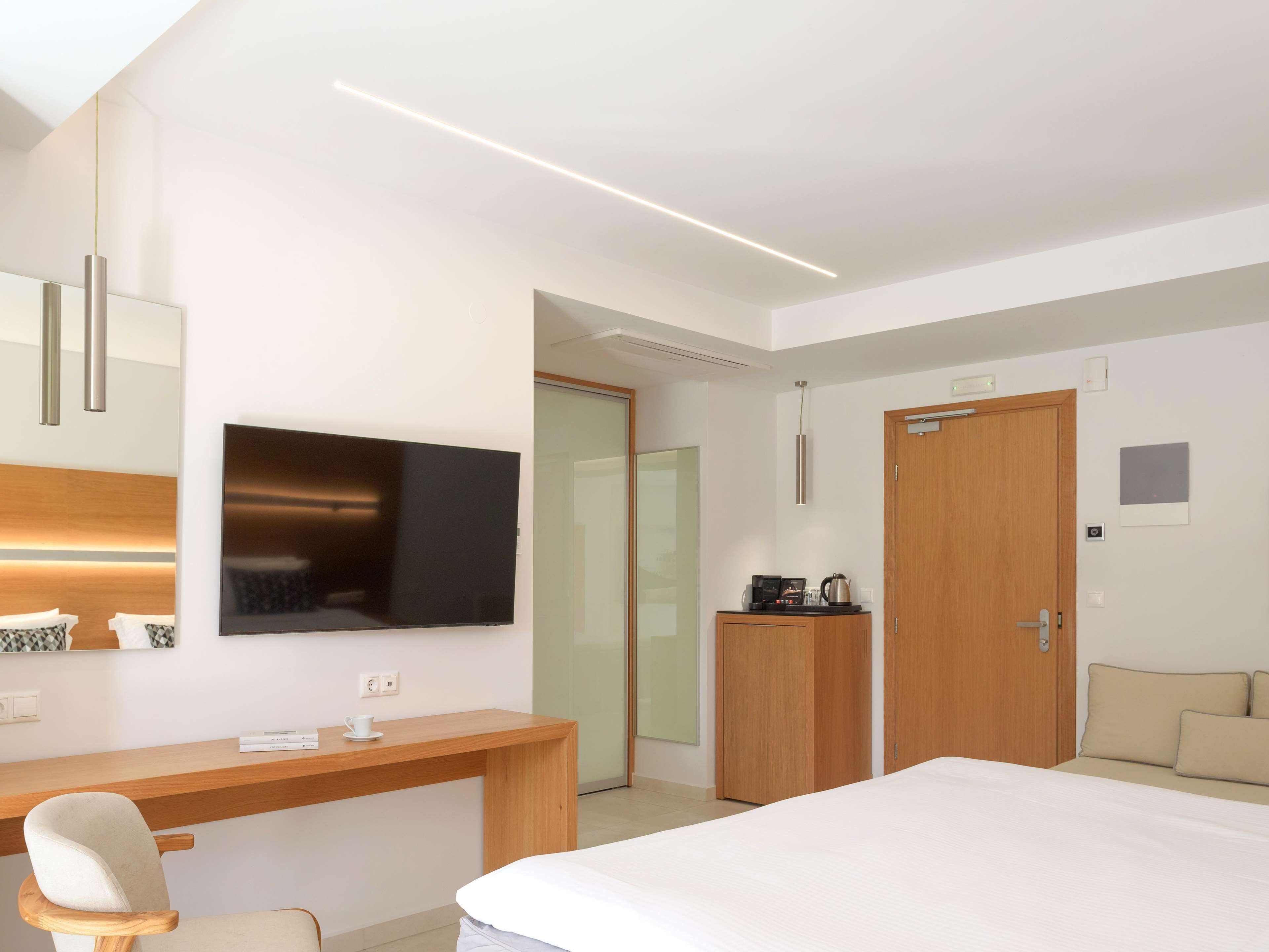 A modern hotel room with a bed, a desk, a chair, a TV, a wardrobe, and a coffee machine.