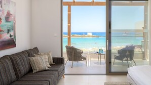 A room with a sofa, a chair, and a bed. There is a glass door leading to a balcony with a view of the sea and a building. - High Beach White (Hersonissos)