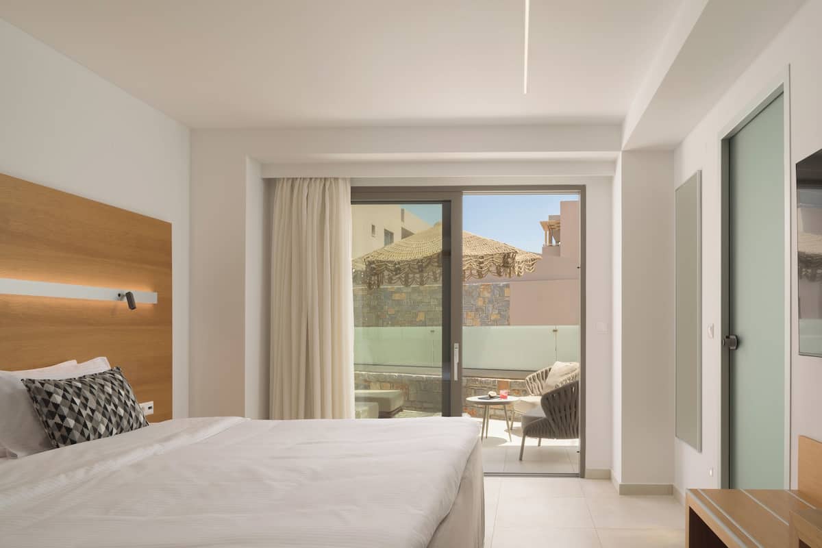 A hotel room with a bed, a small table, a chair, and a balcony with a view of buildings.