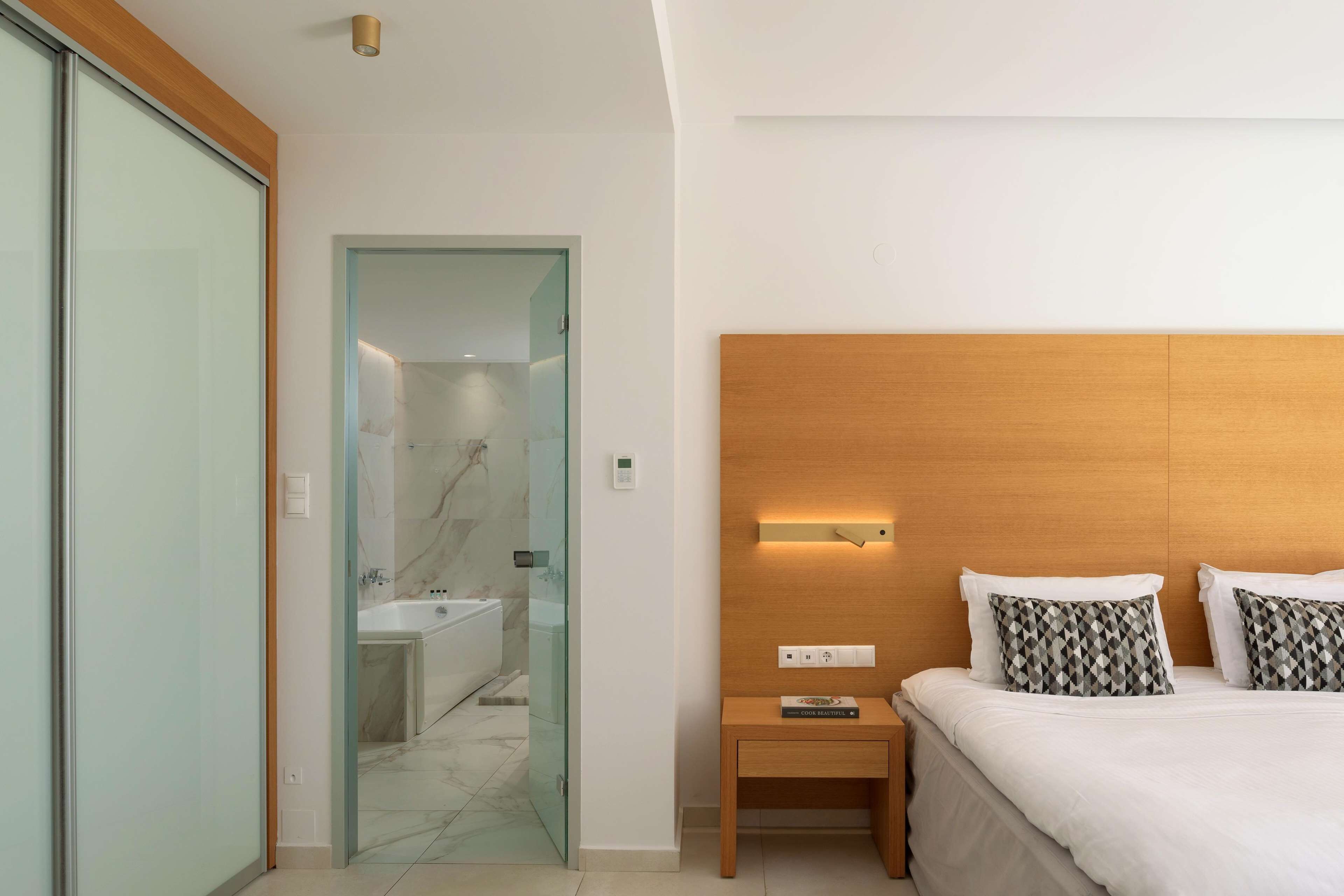 A modern hotel room with a glass door leading to a bathroom, a wooden bedside table with a book, and a neatly made bed with patterned pillows.