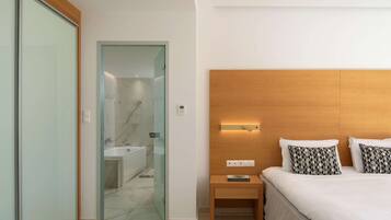 A modern hotel room with a glass door leading to a bathroom, a wooden bedside table with a book, and a neatly made bed with patterned pillows.