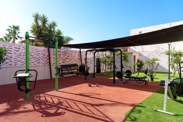 An outdoor gym area with exercise equipment, a red rubberized floor, and a white wall with a geometric pattern.
