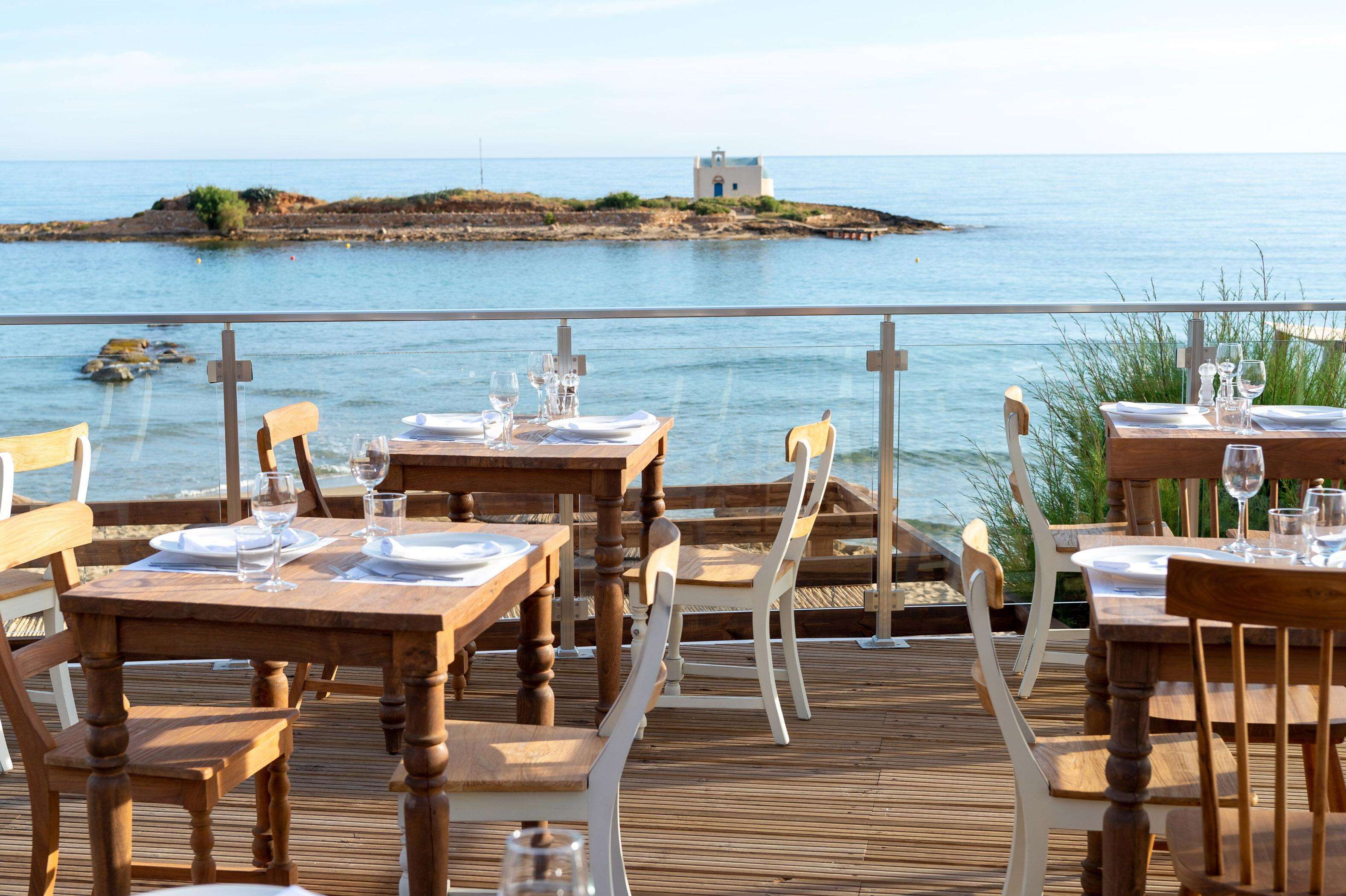 Une terrasse en bois, agrémentée de tables et de chaises pour les repas, offre une vue sur une étendue d’eau avec une petite île et un bâtiment situé au loin.