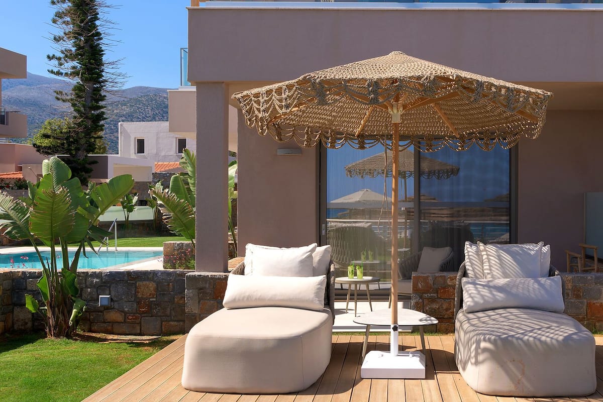 A modern outdoor seating area with two cushioned chairs and a woven umbrella, overlooking a pool and a mountainous landscape.