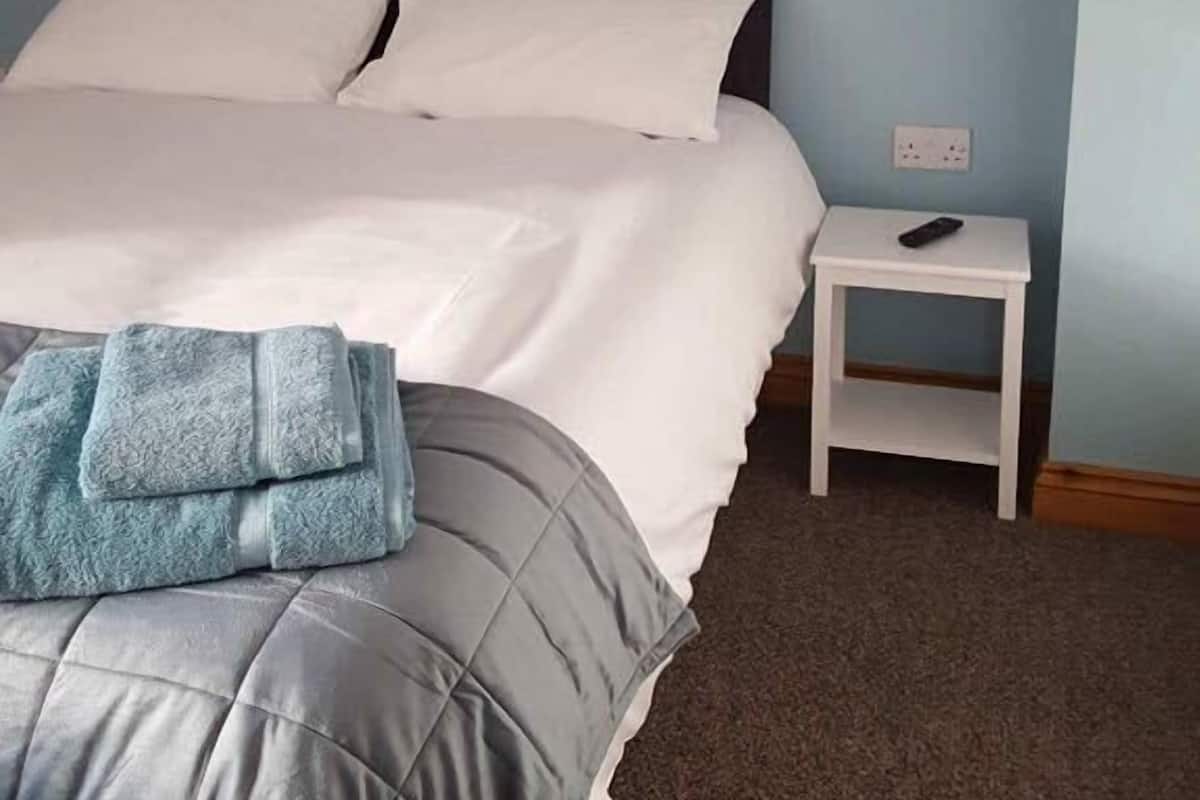 A neatly made bed with white linens and a grey quilt, a small bedside table with a lamp, and a remote control on it.