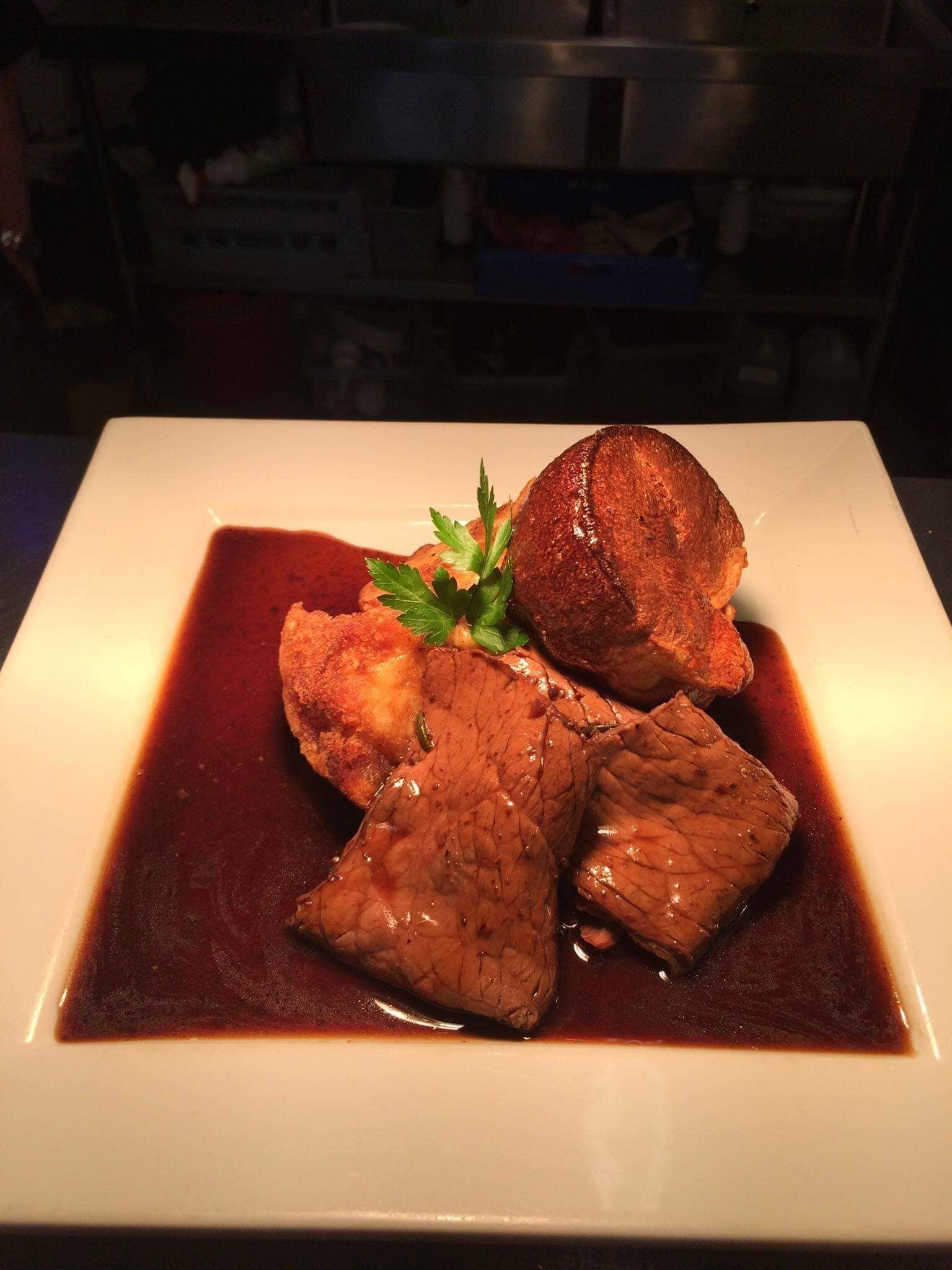 Un plato blanco cuadrado con un trozo de carne y una guarnición sobre un fondo oscuro.