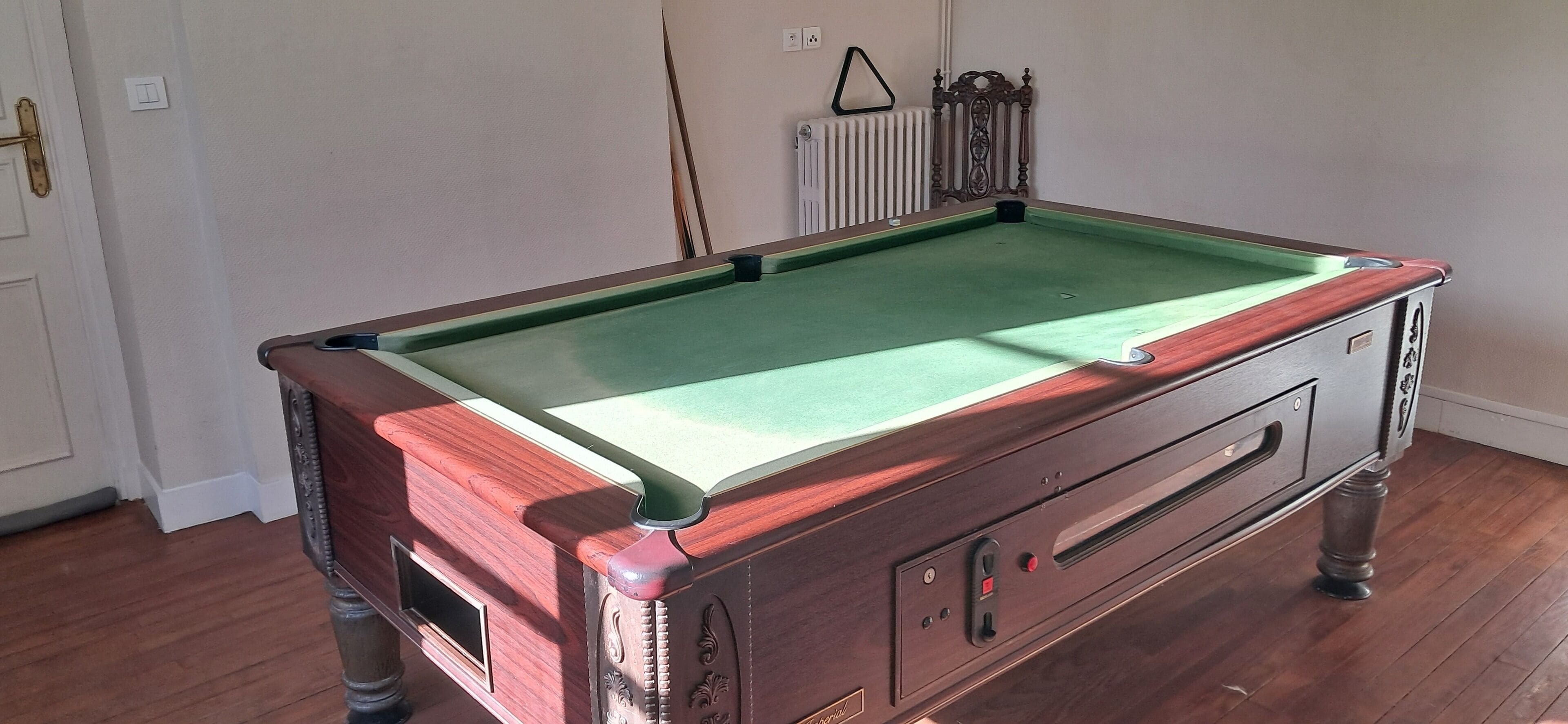 Una mesa de billar con superficie de fieltro verde, ubicada en una sala con piso de madera y un radiador al fondo.