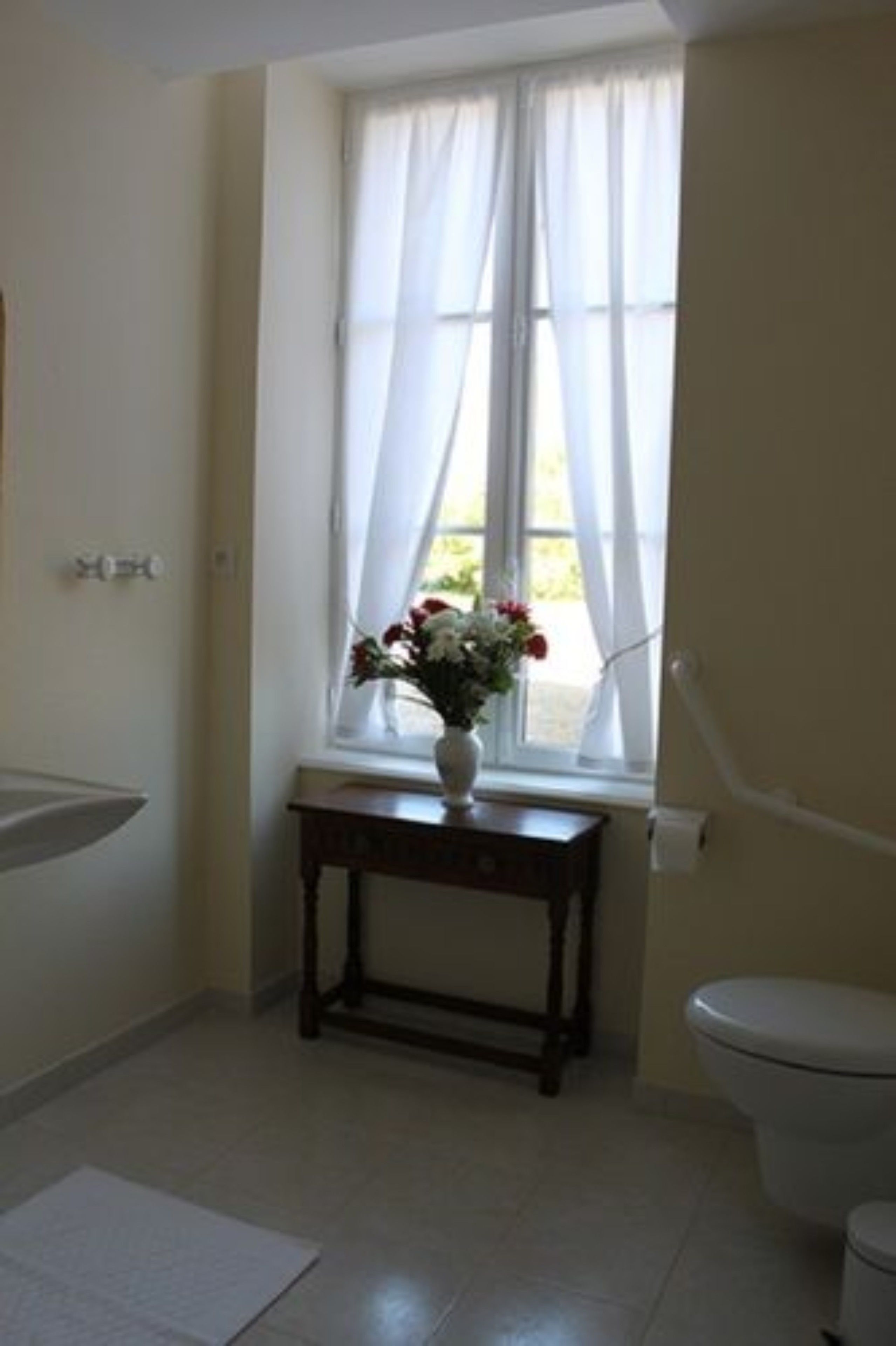 A bathroom with a toilet, a sink, a window with sheer curtains, and a small table with a vase of flowers.