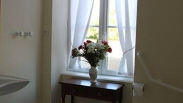 A bathroom with a toilet, a sink, a window with sheer curtains, and a small table with a vase of flowers.