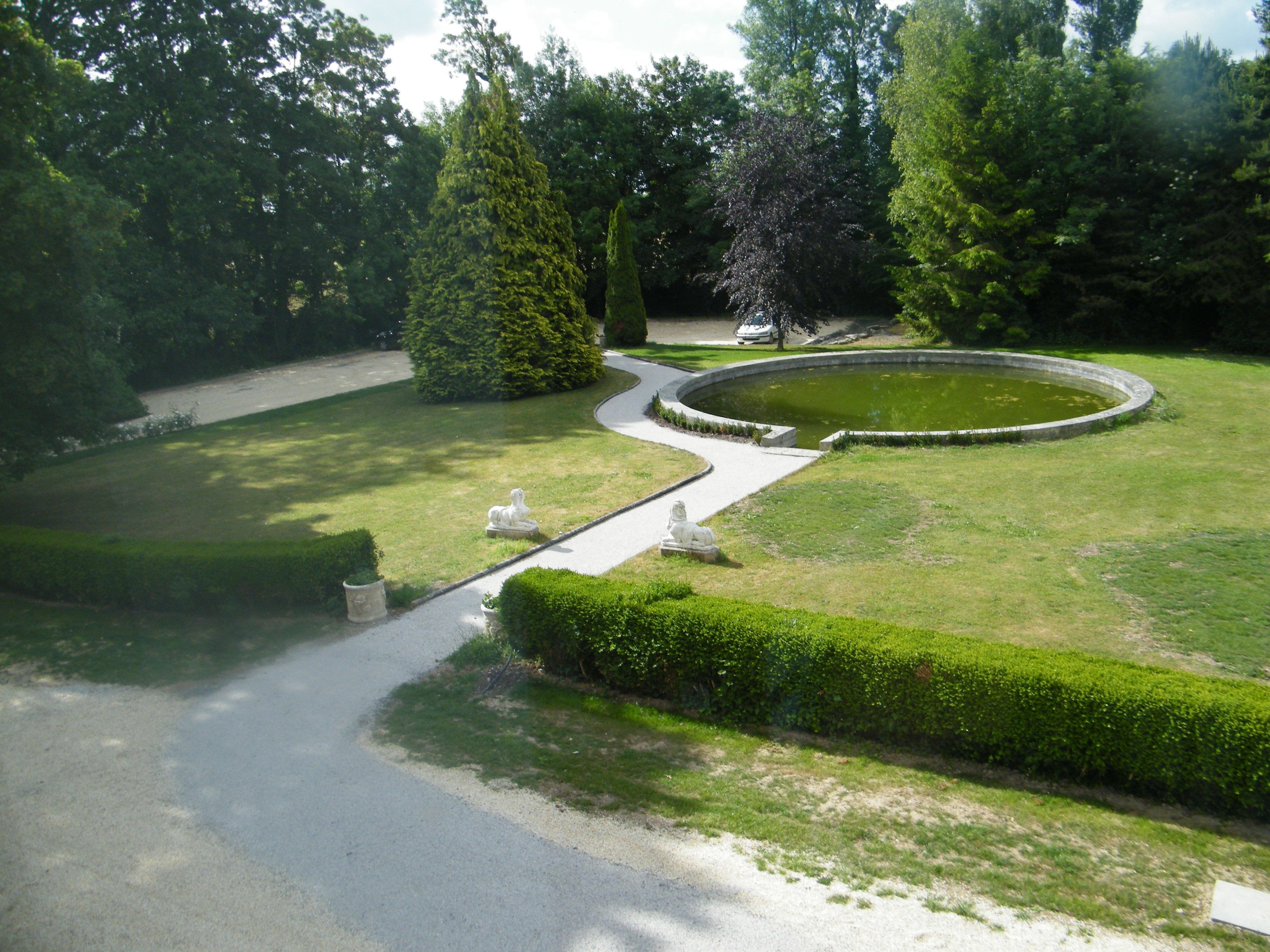 Ein gepflegter Garten mit einem kreisrunden Teich, ordentlich geschnittenen Hecken und einem geschwungenen Weg.