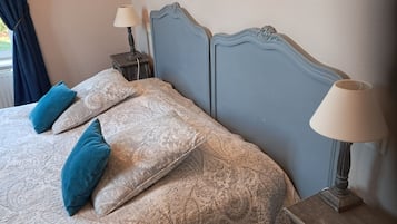 Un llit amb roba de llit estampada i dos coixins, una taula de nit amb una làmpada, i una finestra amb cortines.