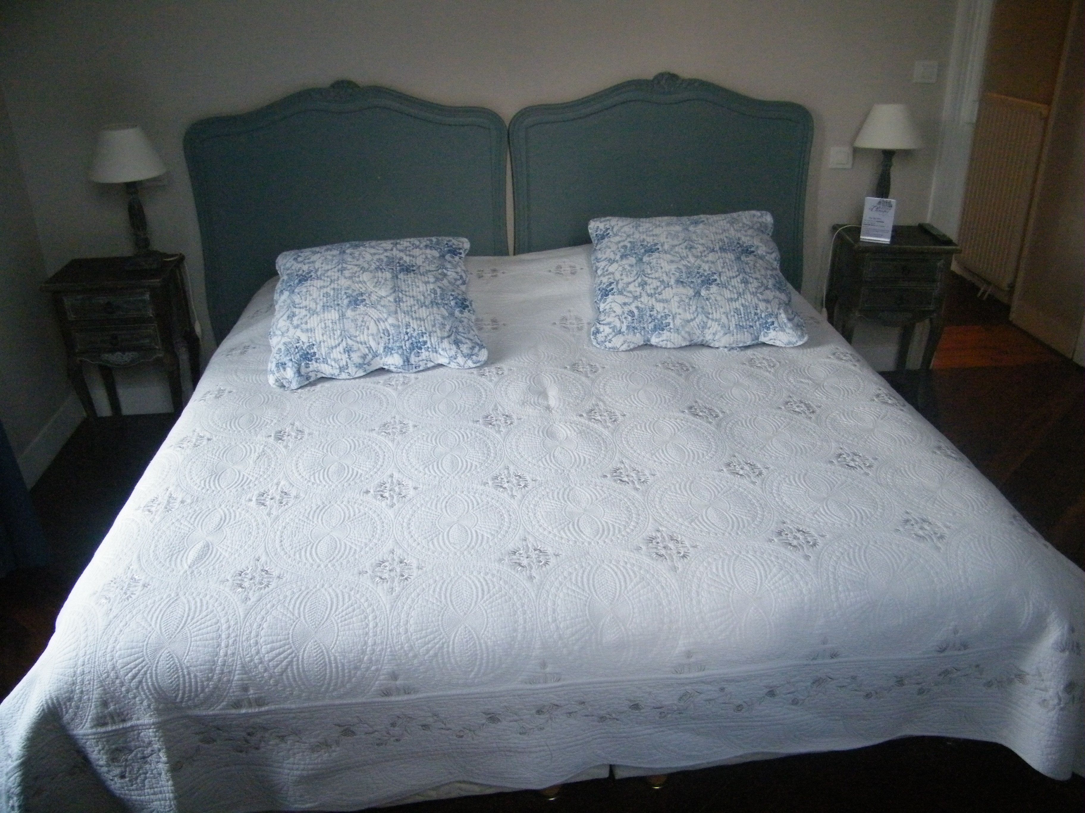 Uma cama de casal com colcha branca e duas almofadas azuis estampadas, com duas mesas de cabeceira e luminárias de cada lado.