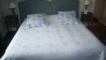 Uma cama de casal com colcha branca e duas almofadas azuis estampadas, com duas mesas de cabeceira e luminárias de cada lado.
